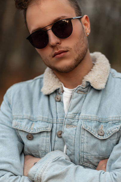Portrait d'un élégant jeune homme hipster en lunettes de soleil dans une veste vintage en denim bleu avec une coiffure tendance à l'extérieur dans un parc. Mannequin attrayant pour une promenade dans la forêt d'automne
. - Photo, image