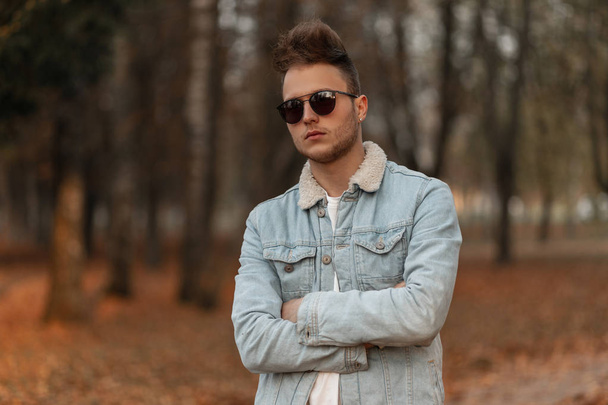 Fashionable young man with a beard with a stylish hairstyle in trendy sunglasses in a blue denim jacket is relaxing outdoors in the park on an October day. Handsome hipster guy. Youth autumn fashion. - Photo, Image