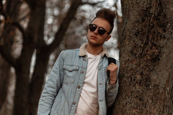 Mladý cool hipster muž ve stylových slunečních brýlích v retro modré džínové bundě s módním účesem v bílém tričku s batohem odpočívá u stromu v parku. Atraktivní chlap venku. - Fotografie, Obrázek