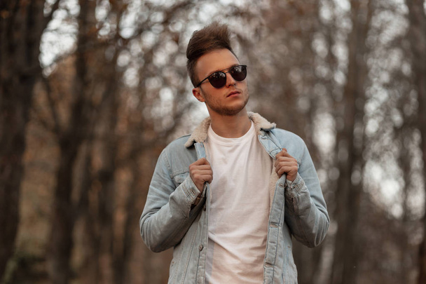 Trendy young man straightens a stylish denim jacket. Handsome hipster guy in black sunglasses with fashionable hairstyle posing in the autumn forest. New collection of men's clothing. Youth look. - Photo, Image