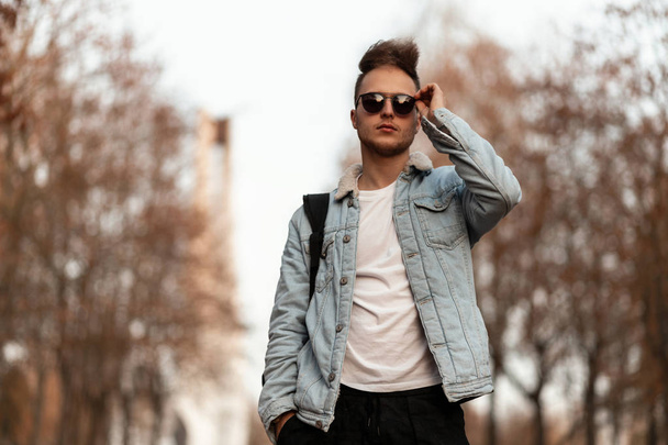 Stylish young man with a beard with a fashionable hairstyle in trendy sunglasses in a blue denim jacket is relaxing outdoors in the park on an October day. Handsome hipster guy. Youth autumn fashion. - Fotografie, Obrázek
