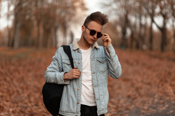 Jovem moderno e moderno com um penteado elegante em óculos de sol pretos em uma jaqueta de ganga com uma mochila posando na floresta de outono. Cara hipster atraente em roupas da moda ao ar livre em uma floresta
. - Foto, Imagem