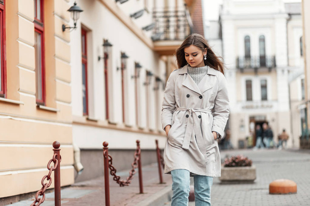 Europejska stylowa młoda kobieta w niebieskich dżinsach w płaszczu modowym spaceruje po mieście w ciepły jesienny dzień. Atrakcyjna dziewczyna w modnych ubraniach na świeżym powietrzu. Kolekcja jesienno-wiosenna odzieży damskiej - Zdjęcie, obraz