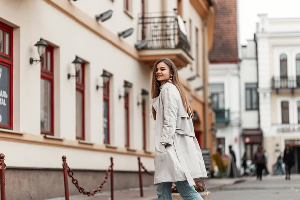 Happy elegant young woman model in a fashionable trench coat with chic brown long hair with natural make-up outdoors in the city. Joyful girl smiling and walks down the street. Spring style.  - Foto, afbeelding