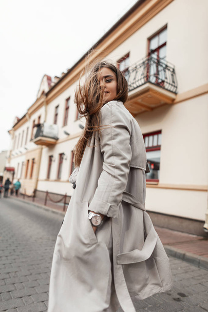 View from the back on a positive attractive young woman in a fashion trench coat with long chic brown hair on a street in the city. Beautiful girl travels through the city streets on a warm autumn day - Foto, immagini
