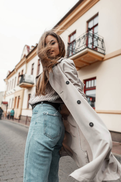 Modern pretty trendy young woman in a warm vintage knitted sweater in an elegant jeans in trench coat posing outdoors on a autumn day. Cute fashionable girl fashion model goes and turns back. - Fotografie, Obrázek