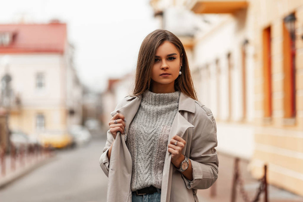 Trendy portrait of a young woman in stylish clothes on the street. Cute girl in a knitted sweater stands and straightens an elegant trench coat outdoors near vintage buildings on a bright spring day. - Zdjęcie, obraz