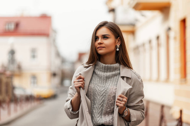 Pretty cute young woman with chic brown long hair with natural make-up in fashionable trench coat in knitted sweater posing in a spring day outdoors. Stylish attractive girl model resting in the city. - Photo, Image