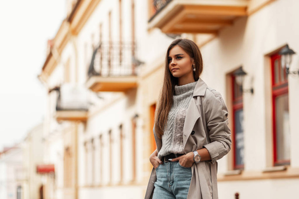 Model of a pretty elegant woman in a chic beige trench coat with brown long hair in a stylish knitted sweater in a jeans walks around the city on a bright spring day. Modern cute girl. Youth fashion. - Photo, Image