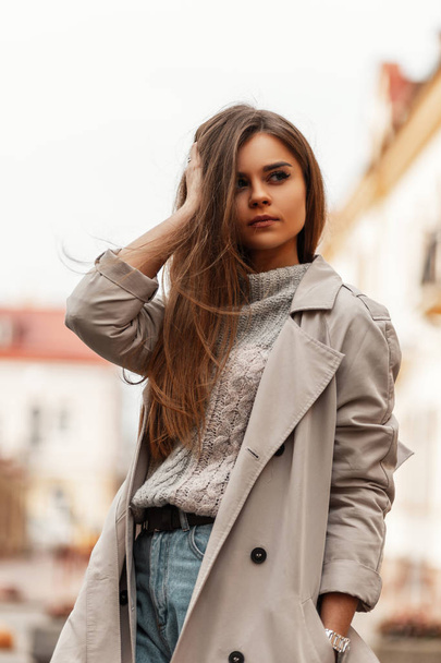 Pretty urban young woman straightens hair. Beautiful stylish girl a fashion model in a fashionable trench coat in a vintage gray knitted sweater walks around the city near the buildings. Spring style. - Foto, immagini