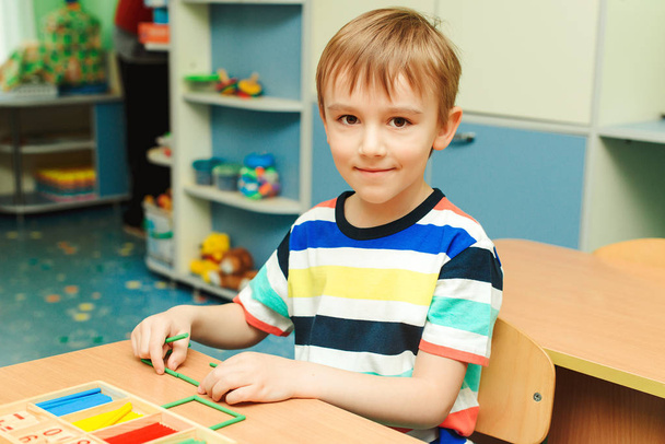 Szczęśliwy chłopak siedzi przy biurku w klasie. Dzieciak używa kolorowych kijów i uczy się liczyć..  - Zdjęcie, obraz