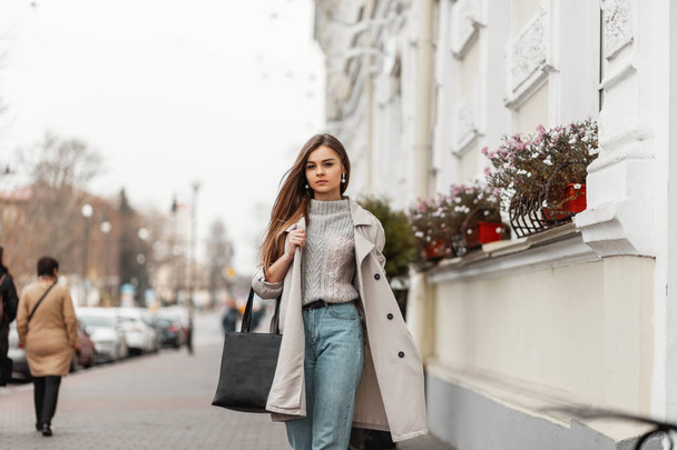 革のヴィンテージバッグを着たファッショナブルなアウターを着たヨーロッパの若い女性が、ファサードに花のある白い建物の近くの通りを歩いています。暖かい秋の日にはスタイリッシュな女の子が街を歩く. - 写真・画像