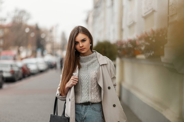 Modern, fiatal barna hajú nő kötött pulóverben, divatos hosszú ballonkabátban, fekete bőrzsákkal a szabadban pózolva egy meleg őszi napon. Elegáns városi lány modell egy utcában a városban. - Fotó, kép