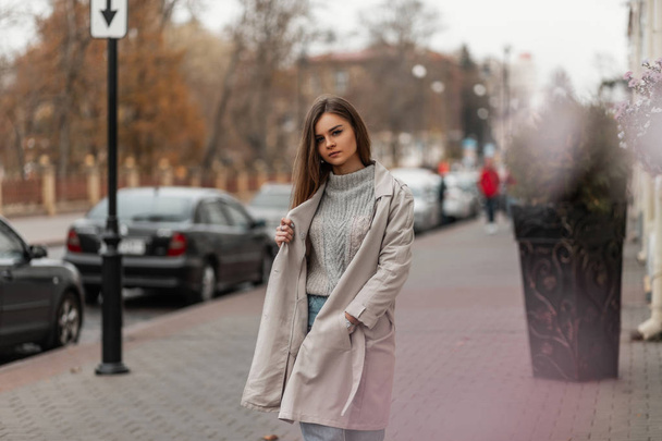 Pretty elegant young woman in a stylish trench coat in a fashionable knitted sweater posing standing on the street near the road. Attractive urban girl is walking on the city on a warm autumn day. - 写真・画像