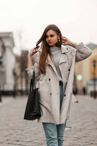 Young beautiful woman in a fashionable long trench coat in blue vintage jeans with a stylish black leather bag enjoy a walk along the street in the city. Attractive trendy brown-haired girl outdoors. - Fotografie, Obrázek