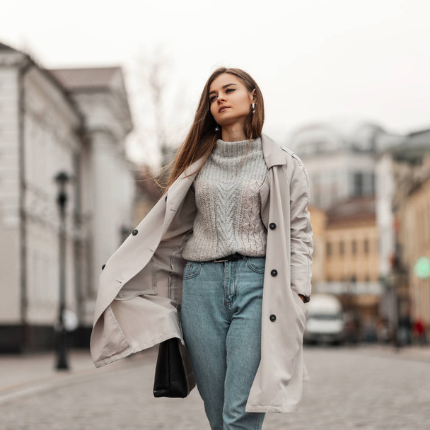 ヨーロッパの若い茶色の髪の女性がトレンディーなジーンズの革袋のニットヴィンテージセーターでエレガントなトレンチコートを着て街を歩きます。魅力的な女の子モデルは通りを旅します. - 写真・画像