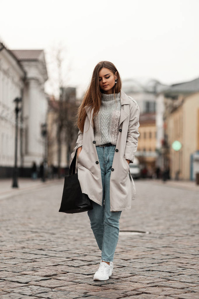 Pretty young woman model in a stylish trench coat in vintage jeans in white trendy shoes with a leather bag in a knitted sweater walks along a stone road in the city. European urban girl enjoys a walk - Photo, Image