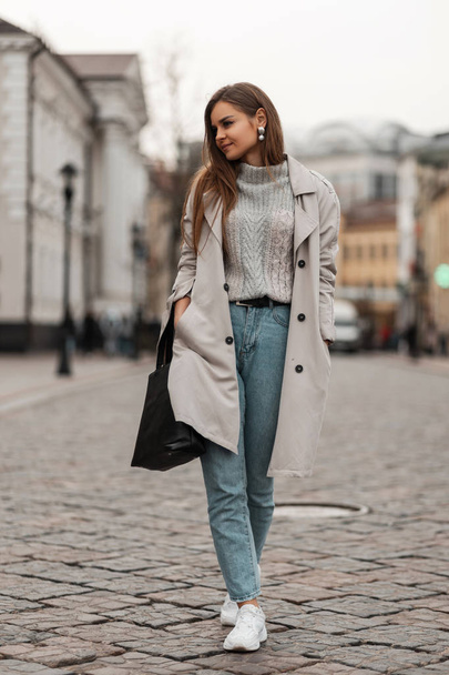 European beautiful young woman in stylish spring-autumn outerwear in sneakers with a fashionable black bag walks along a city street near vintage buildings. Elegant girl model outdoors. Youth fashion. - Fotó, kép