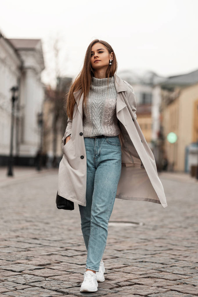 Jeune belle femme dans un pull tricoté à la mode en jeans dans un élégant trench coat avec un sac en cuir noir en baskets marche sur une route en pierre dans la ville. attrayant fille modèle voyages sur la rue
 - Photo, image