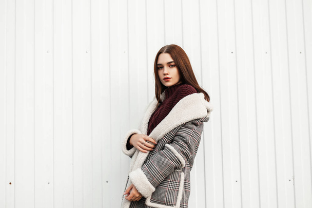 Urban young stylish brown-haired woman in a stylish checkered long jacket with fur in a fashionable burgundy warm sweater stands on the street near a modern white building. Pretty trendy girl outdoors - Foto, immagini