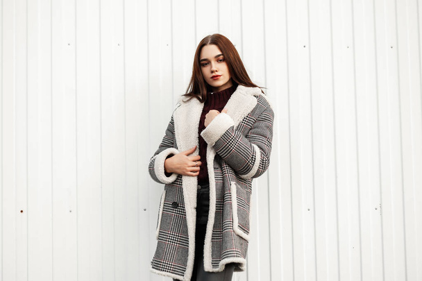 Gyönyörű modell egy vonzó fiatal nő egy vintage kockás kabát szőrme egy elegáns kötött pulóver áll a város közelében egy fehér csíkos fal.Aranyos barna hajú lány pózol az utcán - Fotó, kép
