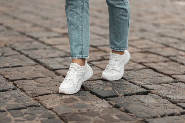 Современная молодая женщина в стильных синих джинсах в модных белых кроссовках стоит на каменной дороге в городе. Стильная женская обувь. Молодежный стиль. Крупный план женских ног в модных туфлях
. - Фото, изображение