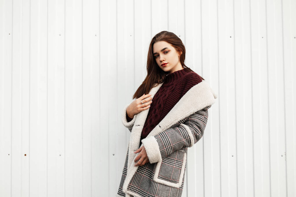 Leuke jonge bruinharige vrouw in een trendy warme geruite jas met bont in een stijlvolle gebreide trui poseert bij de witte muur. Aantrekkelijk meisje staat in stijlvolle kleren op een witte achtergrond. - Foto, afbeelding
