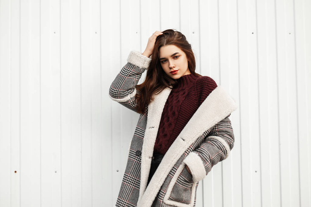 Young beautiful woman fashion model in trendy clothes on a white background. Attractive girl in a fashionable jacket with fur in a knitted sweater posing in the city near a modern striped wall.  - Photo, Image