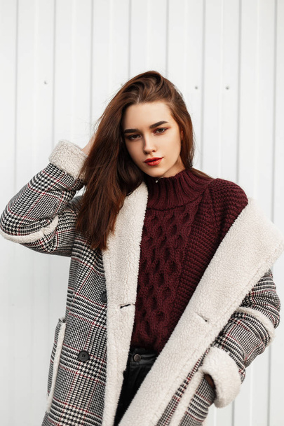 Model van een aantrekkelijke jonge vrouw in een stijlvolle gebreide bordeaux trui in een geruite warme lange jas met bont poserend bij een witte muur op straat. Mooi modieus meisje buiten. - Foto, afbeelding