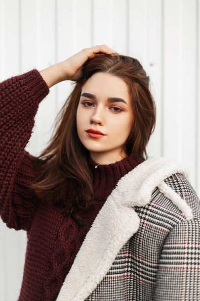 Pretty beautiful portrait of a cute young woman in a vintage burgundy sweater in a fashionable jacket with natural make-up on a white background outdoors. European modern girl posing in the street. - Photo, Image