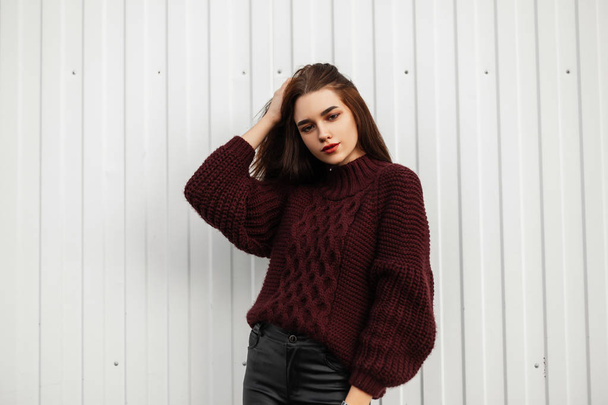 Stylish pretty young urban woman in a fashion burgundy sweater in trendy black leather pants posing in the city near a white wall. Attractive cute girl model outdoors. Youth fashionable autumn clothes - Photo, Image