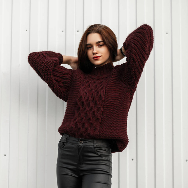 Beautiful European young woman in fashion clothes straightens hair. Urban pretty trendy girl model in a vintage knitted sweater in leather trendy pants posing in the city near a white building. - Photo, Image