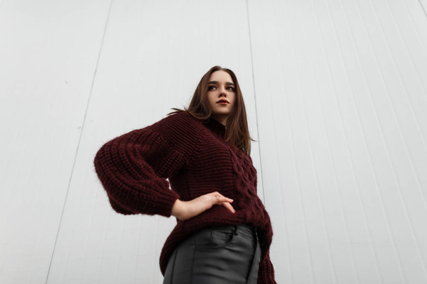 Model of a trendy young woman in a stylish burgundy sweater in leather trendy black pants posing on the street near a white building. Fashionable pretty urban girl in the city on a autumn day. - Photo, Image