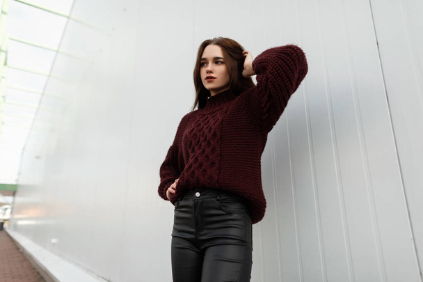 Modell egy divatos fiatal nő egy elegáns burgundi pulóver bőr trendi fekete nadrág pózol az utcán, közel egy fehér épület. Modern szép városi lány a városban egy őszi napon. - Fotó, kép