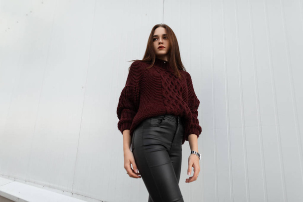 Attractive young woman in a fashionable knitted sweater in stylish leather black pants poses near a modern white wallin the city.Cute european girl fashion model stands on the street near the building - Foto, afbeelding