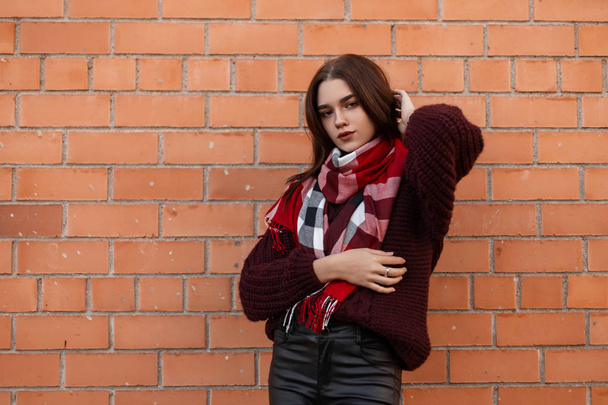 ファッショナブルなバーガンディニットセーターの若いスタイリッシュな女性は、レンガ造りの建物の近くに屋外でポーズをとる革のパンツで赤いトレンディーなスカーフでセーターを編んだ。かわいい美しい女の子は街でリラックスします。秋のファッション. - 写真・画像