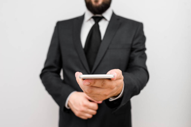 Male human with beard wear formal working clothes hold high technology smartphone device. Man dressed in work suit plus tie holding small mobile hi tech phone using one hand - Photo, Image