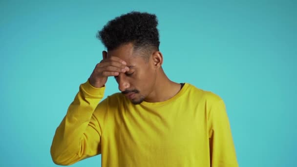 Young student man with afro hair having headache, studio portrait. Guy putting hands on head, isolated on blue background. Concept of problems and headache. - Πλάνα, βίντεο