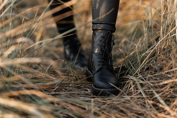 Gros plan des jambes féminines en bottes noires en cuir à la mode. Fille à la mode dans des leggings élégants dans des chaussures de mode marche sur l'herbe sèche dans la rue. Style d'automne décontracté
. - Photo, image