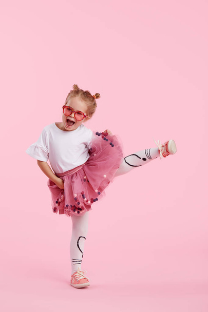 Happy childhood. Funny child girl in tulle skirt jumping and having fun isolated on pink background. Celebrating a vibrant carnival for kids, birthday party. True emotions. Space for text - Фото, изображение