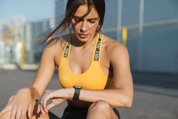 Image of woman looking at smartwatch while sitting on ground out - Foto, Imagen