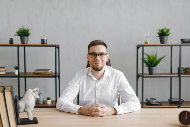 Portrait of young man sitting at his desk in the office and smiling at camera while sitting at workplace - Фото, изображение