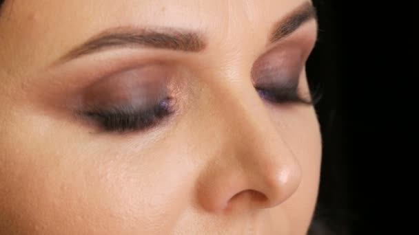 Young beautiful woman with bright blue eyes and evening make-up smoky eyes with blue and purple eyeshadow on black background - Footage, Video
