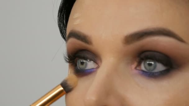 Close-up zicht van een stylist make-up kunstenaar brengt foundation cream met een speciale borstel op het gezicht van een jonge mooie vrouw met blauwe ogen - Video