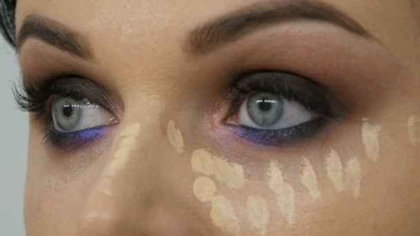 スタイリストメイクアーティストのクローズアップは、青い目の若い女性の顔に特別なブラシでファンデーションクリームを適用します。 - 映像、動画