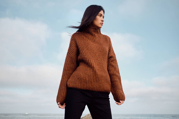 Beautiful stylish Asian girl in knitted sweater thoughtfully looking away on seaside - Photo, Image