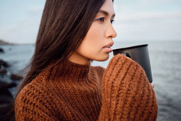 Side näkymä melko aasialainen brunette tyttö neulotut villapaita muki huomaavaisesti sivuun meren rannalla
 - Valokuva, kuva