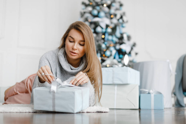 Beautiful girl near the Christmas tree is surrounded by gifts, unpacks gifts - Photo, Image