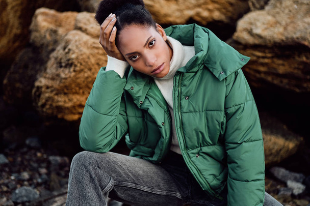 Portrait of casual African American girl in stylish down jacket intently looking in camera posing on stones by the sea - Fotografie, Obrázek