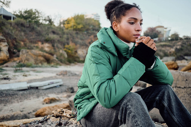 魅力的なスタイリッシュなアフリカ系アメリカ人の女の子でダウンジャケット思慮深く手に座っている石の上に海 - 写真・画像